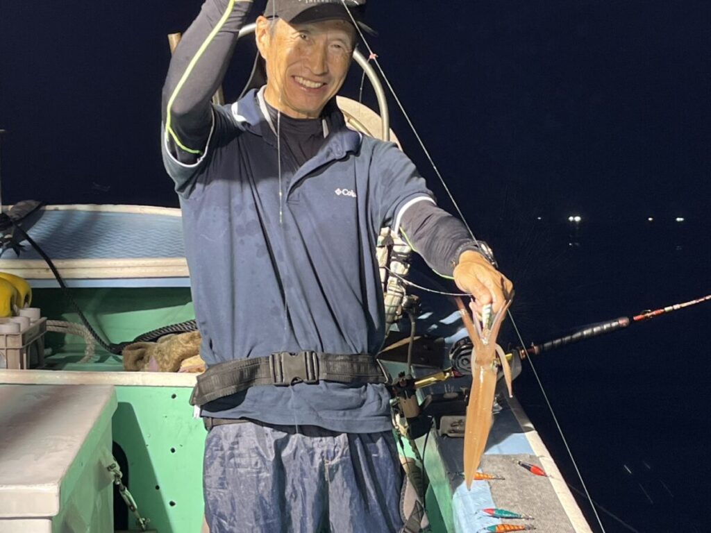 夜、ボートの上に立つ人物が、釣ったイカを釣り糸につけたまま、笑顔で立っている。ボートの上には、さまざまな釣り道具や釣り竿が見える。.