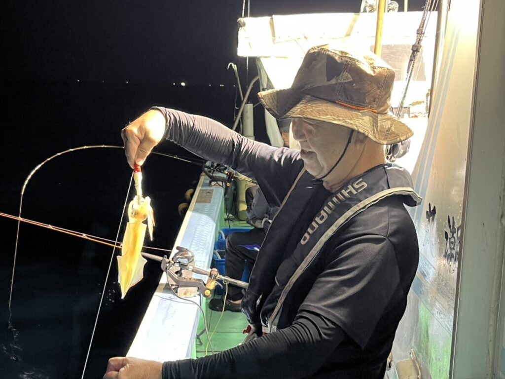 夜、人工の明かりに照らされた船の上で、帽子と釣り道具を身につけた男性が、釣り上げたばかりのイカを掲げている。.