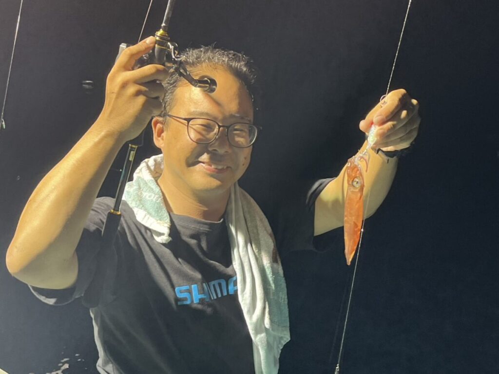 眼鏡をかけた男性が、片手に釣り竿、もう片方の手に小さなイカを持ち、肩にタオルを掛けたまま、暗い背景を背に立って微笑んでいる。.