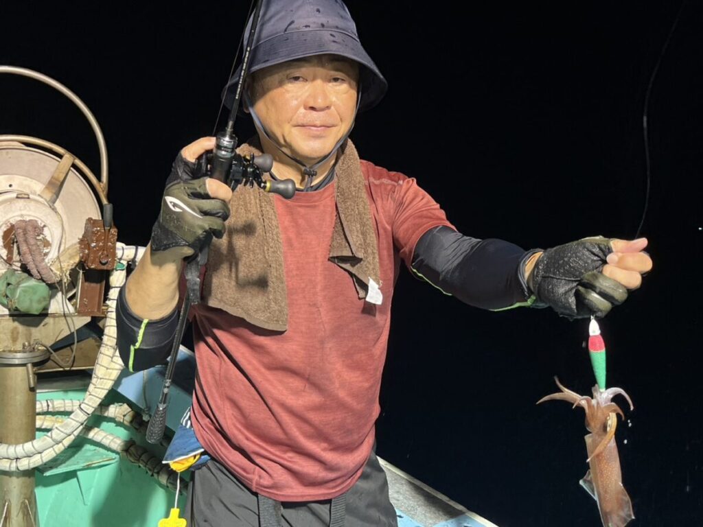 夜、ボートの上に立つ、帽子と手袋を身につけた人物が、釣り竿を手にし、釣り針にかかったイカを見せている。.