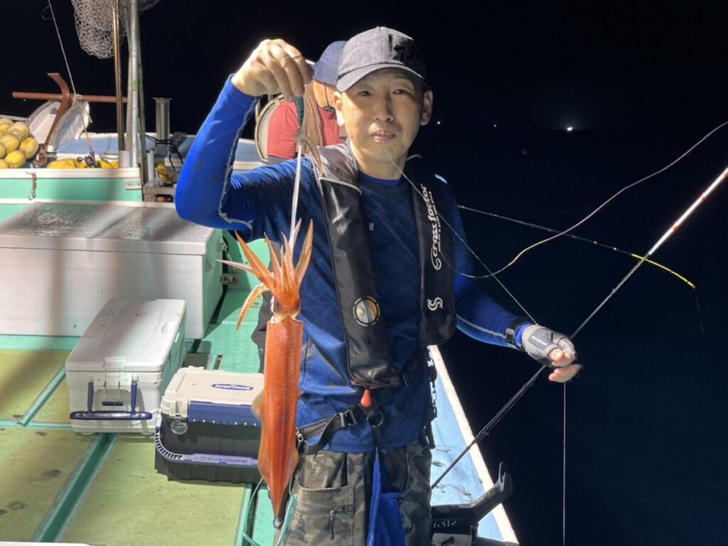 夜、ボートに乗った人物が、釣りで捕まえた大きなオレンジ色のイカを掲げており、周囲には釣り道具や装備が見える。.