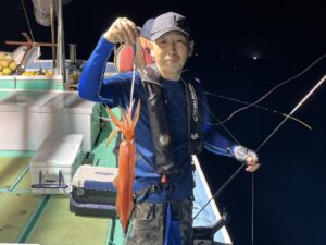 夜、ボートに乗った人物が、釣りで捕まえた大きなオレンジ色のイカを掲げており、周囲には釣り道具や装備が見える。.