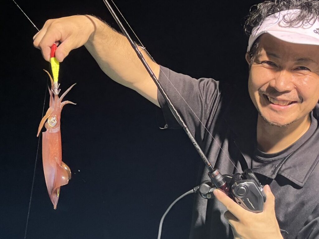 ある人が、夜、釣り竿を手に持ち、釣れたばかりのイカを見せびらかしながら微笑んでいる。.