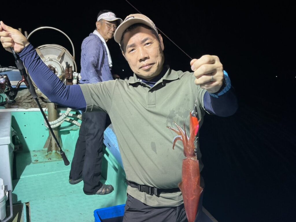 夜、ボートに乗った人物が、獲れたてのイカを掲げている。背景には別の人物が立っている。ボートの甲板はターコイズ色で、釣り道具が見える。.