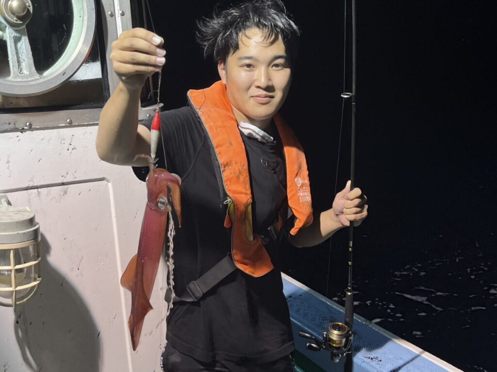 夜、救命胴衣を着た人がボートの上に立ち、片手に釣り竿、もう片方の手には釣ったばかりのイカを持っている。.