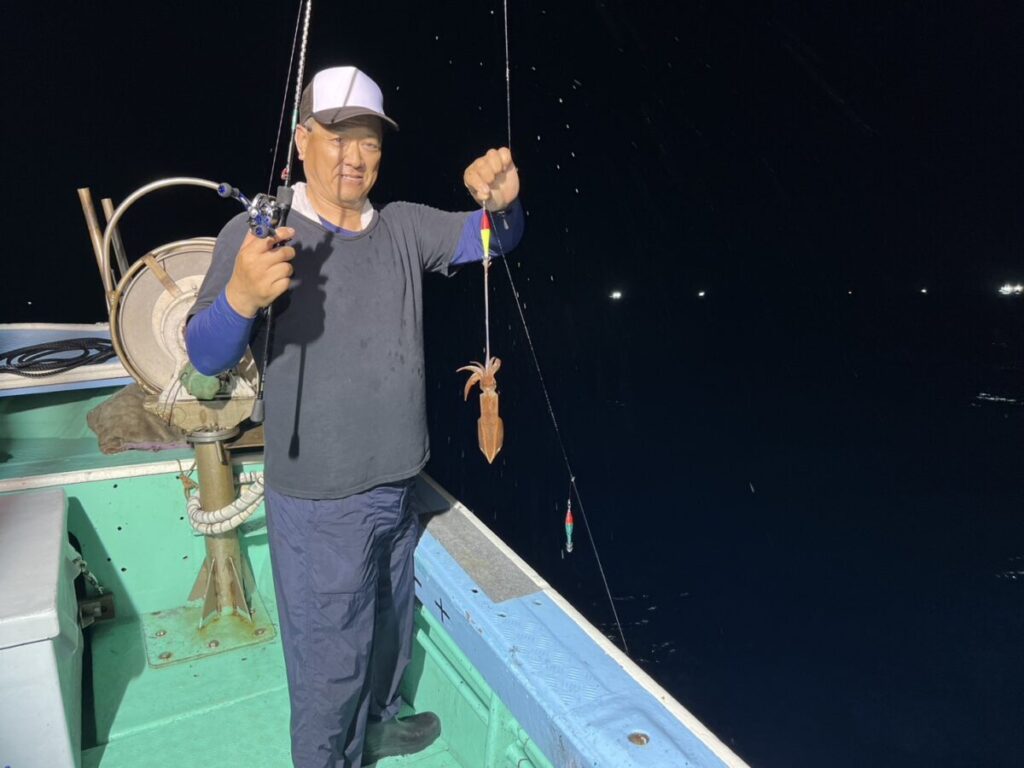 白い帽子と濃い色のシャツを着た人物が、夜、ボートの上に立ち、釣り糸に小さなイカが掛かった釣り竿を手にしている。.