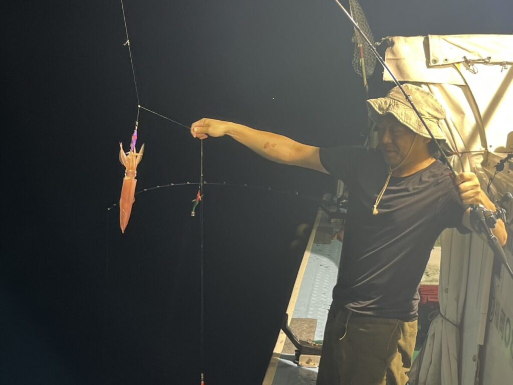 スッテに掛かった赤いケンサキイカを吊り上げて見せる釣り客。玄界灘・宗像大島沖の夜焚きイカ釣り